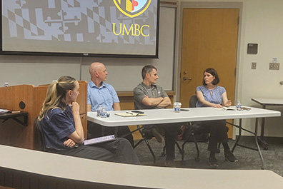 A panel of four people sitting at a long white table during a discussion at UMBC. A large screen behind them displays the university logo.
