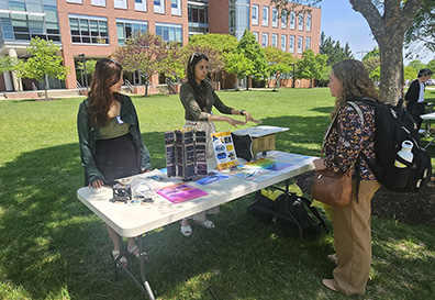 Two women standing behind an outdoor table display with scientific equipment and posters, talking to a visitor on a sunny day.