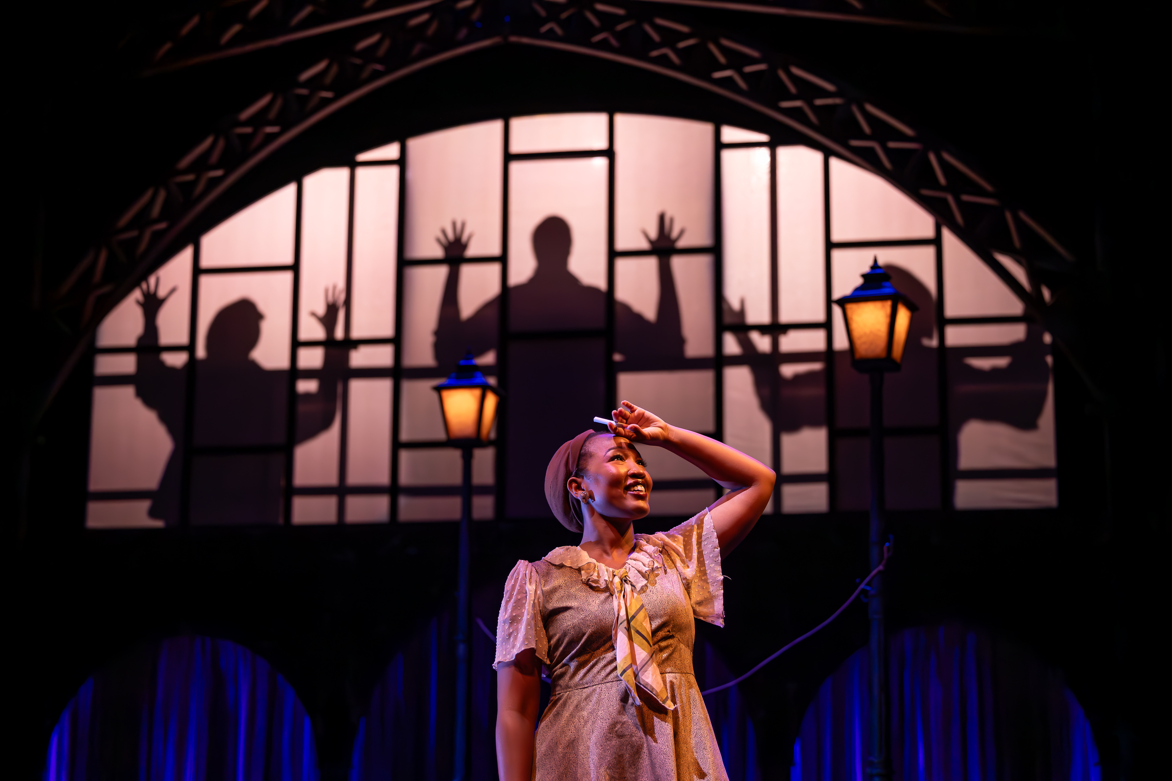 Production Photo from Shakespeare in Harlem. Lulu brushes the sweat off of her forehead with a cigarette in hand. Behind her, we see the shadows of the male ensemble.
