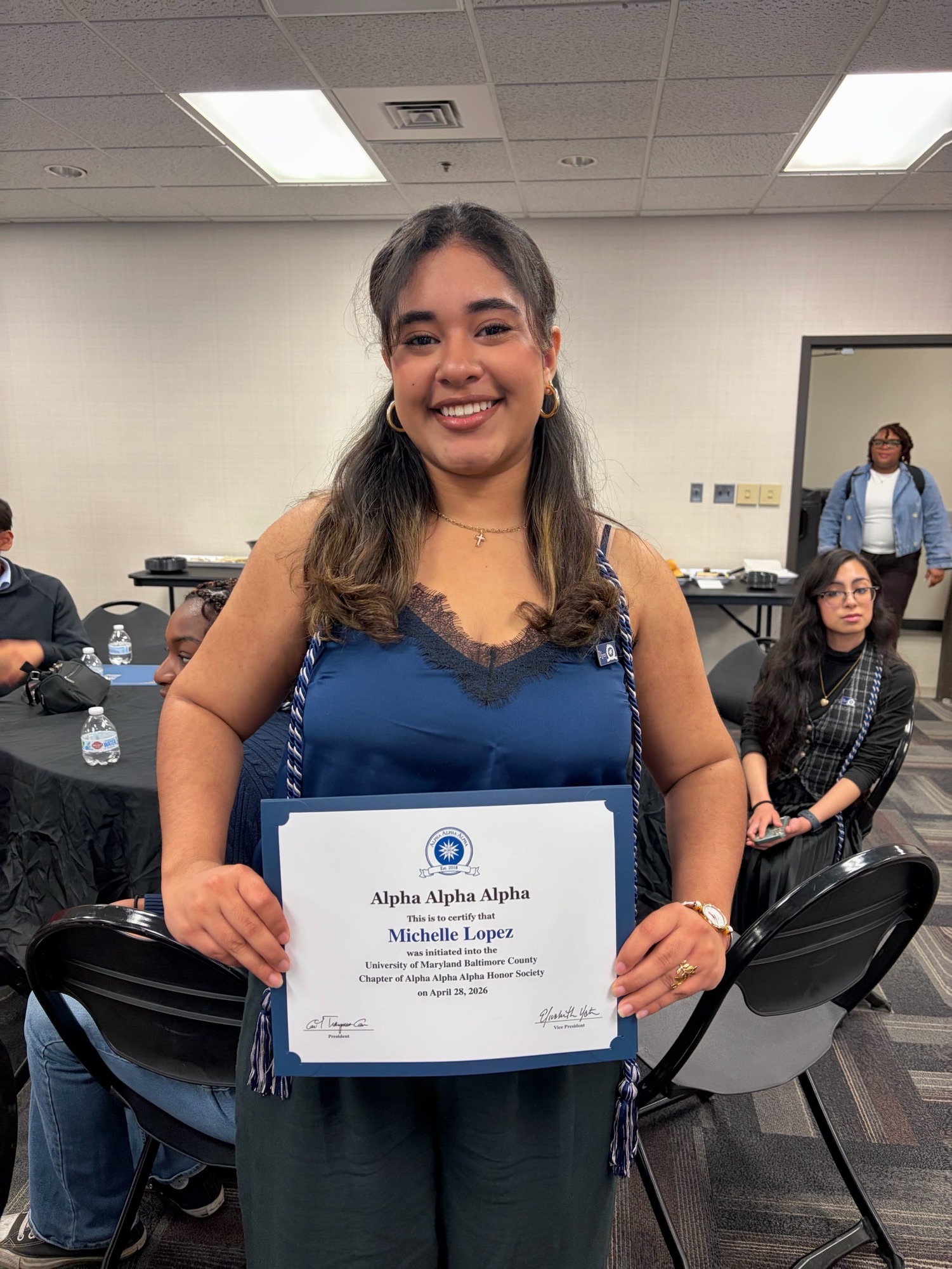 Michelle standing in a room with round tables in a blue top with a blue and whit cord around neck hanging in front of her holding a certificate that says "Alpha Alpha Alpha"