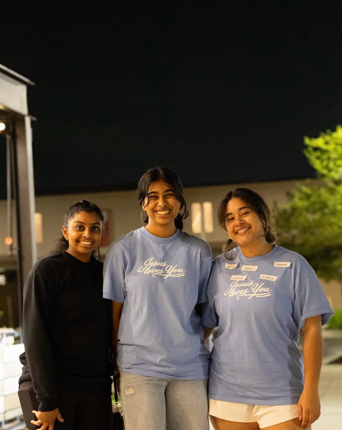 Michelle standing with two students, Michelle and the student next to Michelle has on blue I Love Jesus shirts and the other student has on a black top, all are smiling while standing outside and it looks like nighttime