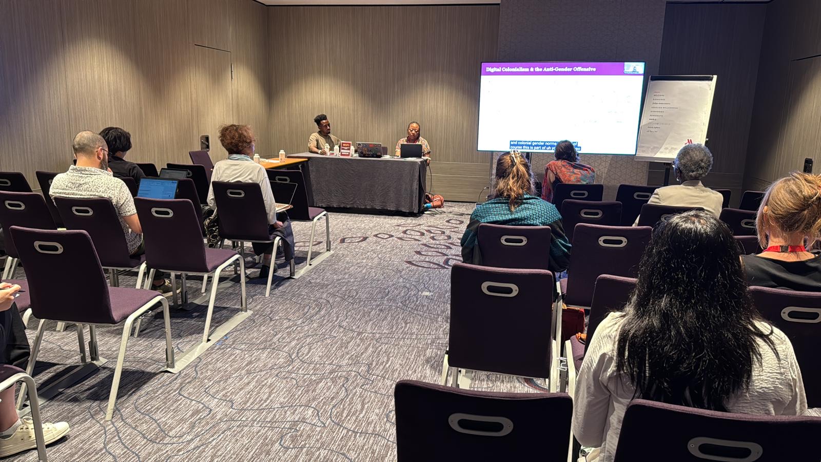 Group of people in conference room, facing front of room. People at panel in front of the room, presentation screen at the front of the room