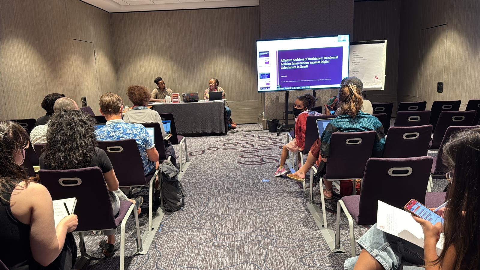 Group of people in conference room, facing front of room. Panel guests at front of room, presentation screen at the front of the room.