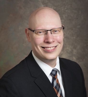 Matthew Trevett-Smith smiling and wearing a black suit and tie. He wears a pair of black-framed glasses.